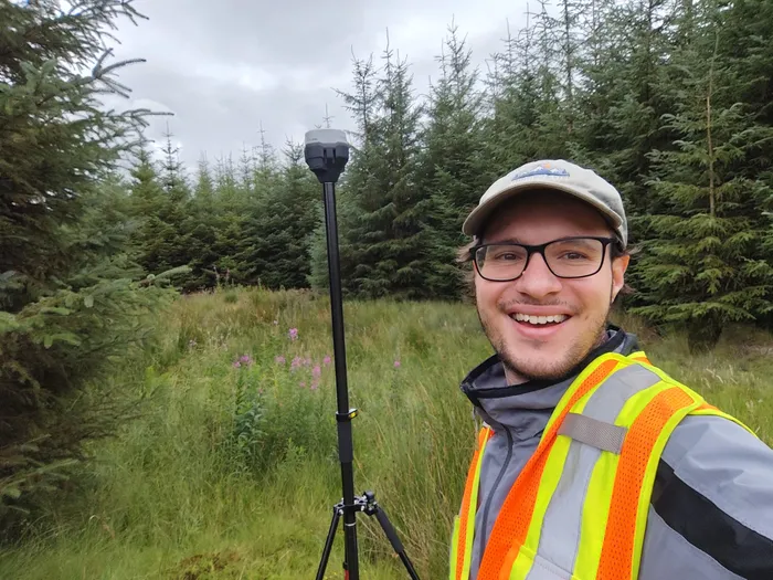 Leonluca Klein smiling in a spruce forest, standing next to a GNSS survey pole on a tripod.