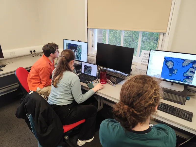 Students and a tutor gathered around a desk of monitors showing maps.