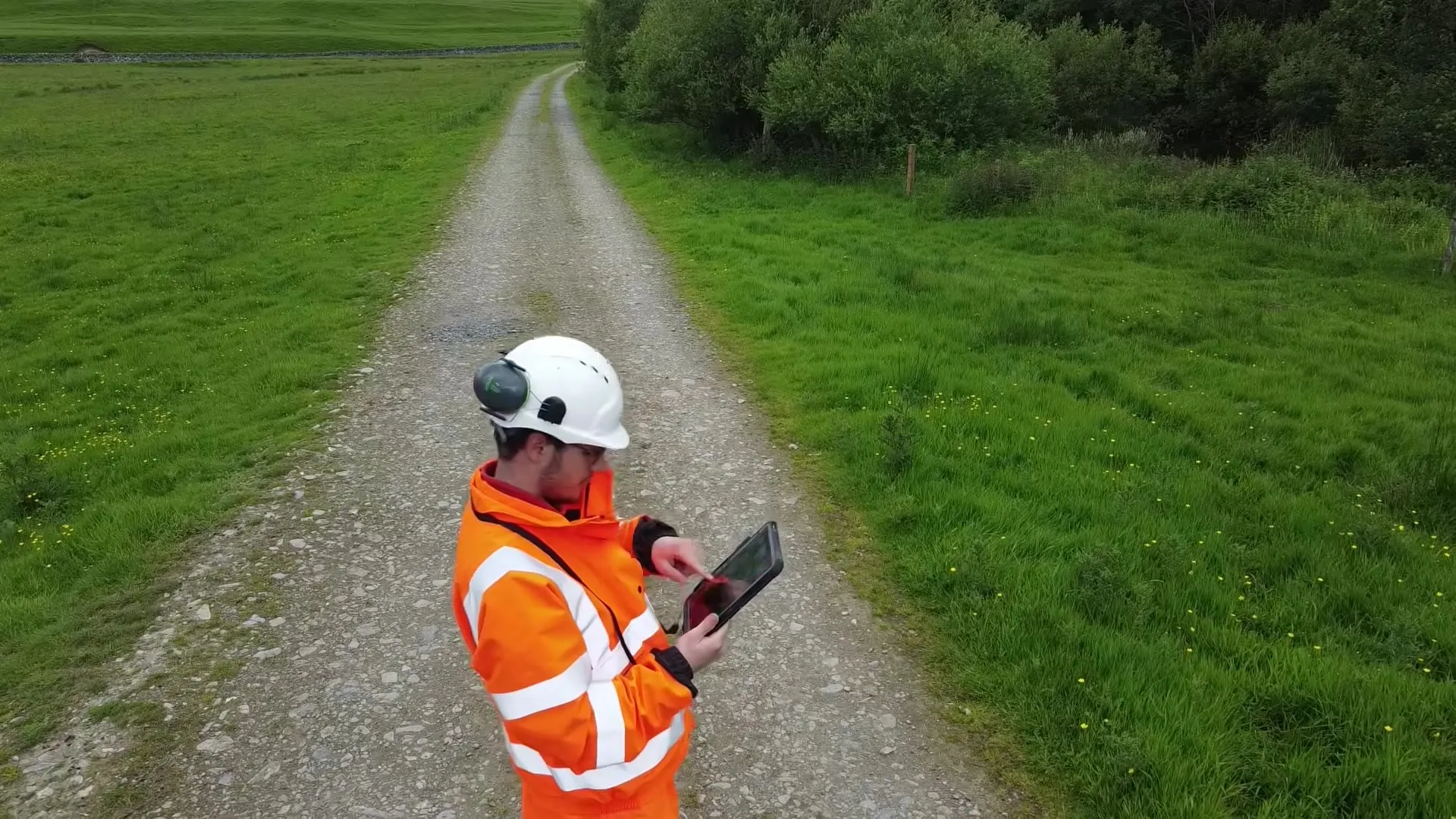 A surveyor in orange hi-vis holding a tablet and pointing along a gravel track.